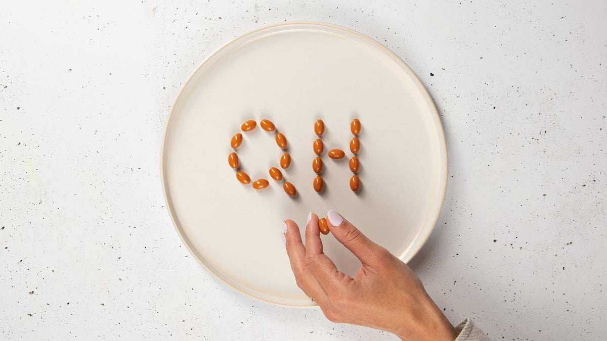 Auf einem Keramikteller sind mit Soft-Gel-Kapseln die Buchstaben Q und H abgebildet, was das Bild QH ergibt. Finger halten eine Soft-Gel-Kapsel in der Hand.