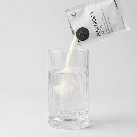 Das geöffnete Sachet von Elektrolyt Aktiv. Das Pulver fällt in ein Glas Wasser.