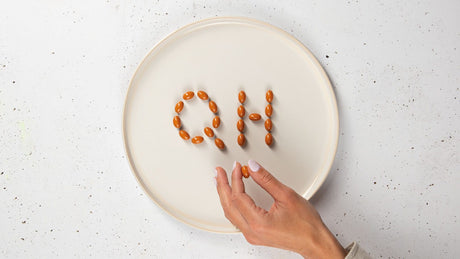 Auf einem Keramikteller sind mit Soft-Gel-Kapseln die Buchstaben Q und H abgebildet, was das Bild QH ergibt. Finger halten eine Soft-Gel-Kapsel in der Hand.
