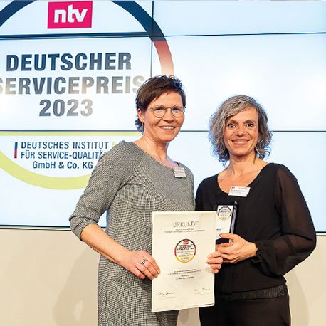 Zwei Frauen halten bei der Verleihung des Deutschen Servicepreises 2023 eine Auszeichnung und eine Urkunde und lächeln in die Kamera.