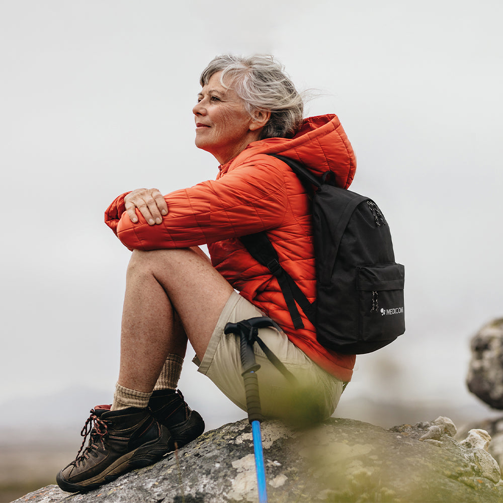 Eine Frau in einer orangefarbenen Jacke mit einem schwarzen Medicom-Rucksack sitzt mit Wanderstöcken auf einem Felsen.