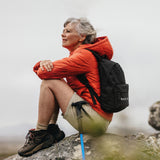 Eine Frau in einer orangefarbenen Jacke mit einem schwarzen Medicom-Rucksack sitzt mit Wanderstöcken auf einem Felsen.